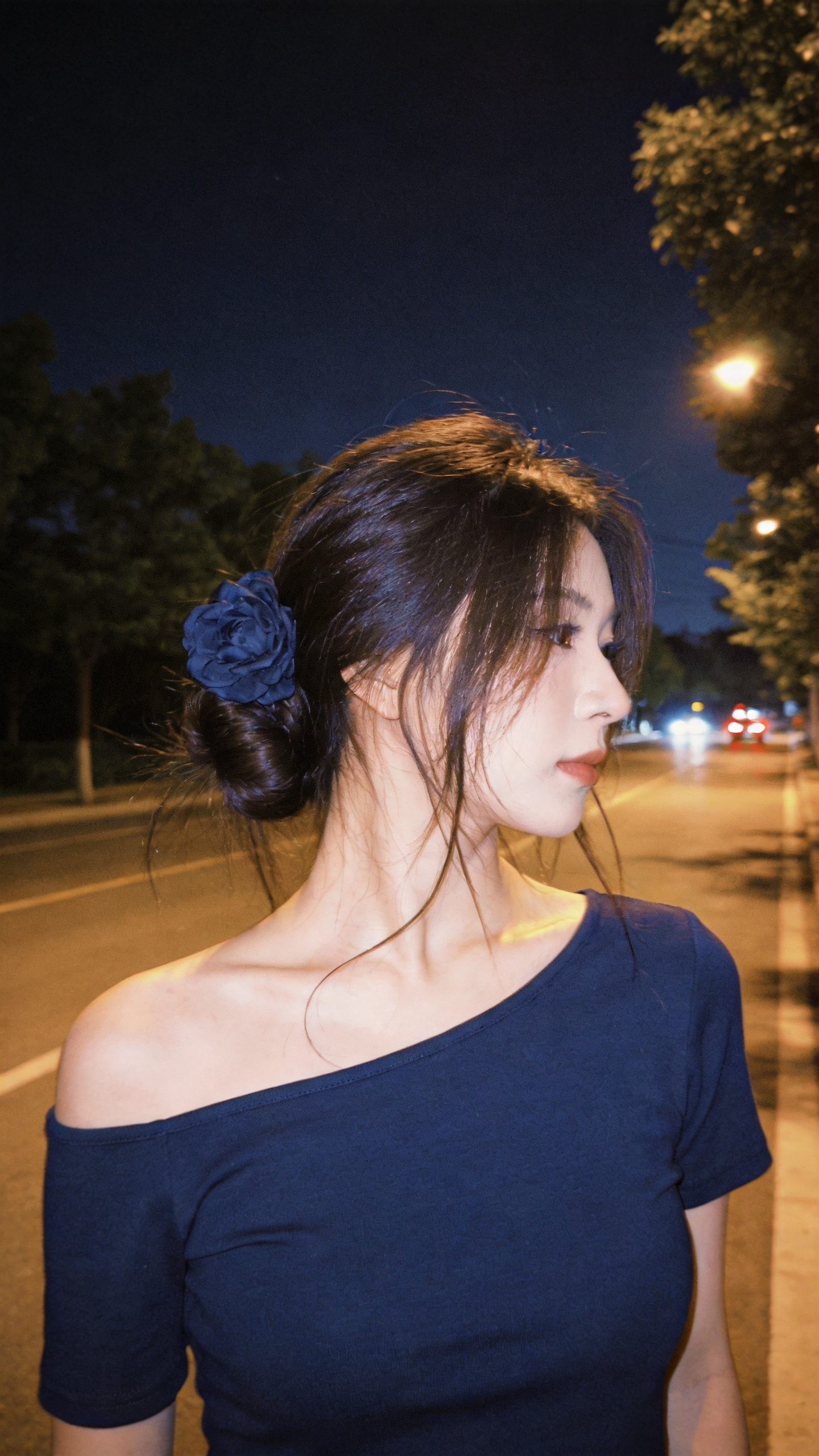 年轻女性，深棕色头发（打理成低发髻，发丝有自然散落的碎发，一侧用深蓝色花朵发饰点缀）；身着深蓝色露肩上衣（面料柔软，剪裁贴合，露出优美肩颈线条）；背景是夜晚的街道（有树木、路灯，远处可见车辆灯光，光线柔和，营造出静谧的… - AI提示词参考图，来自西西提示词