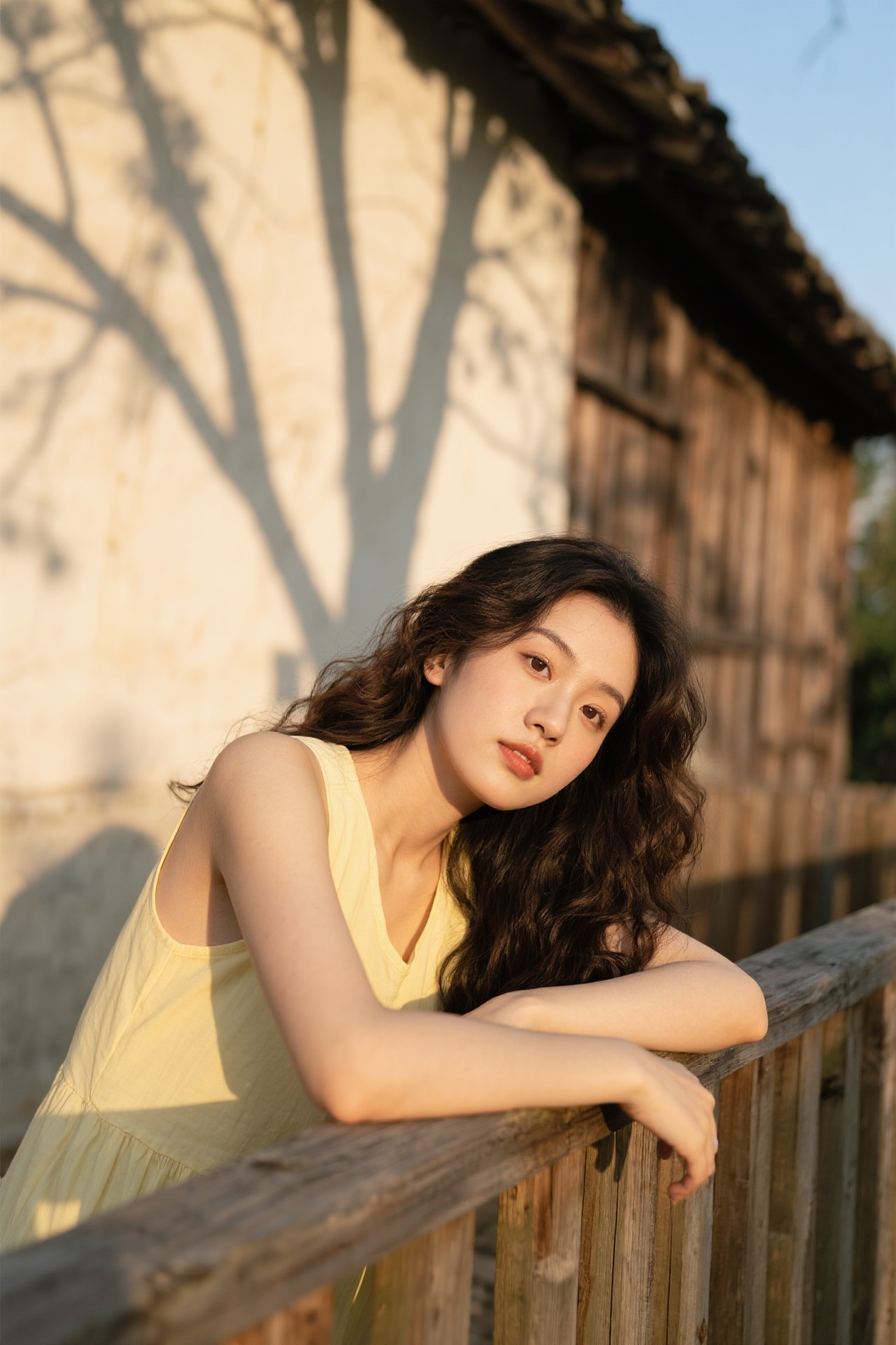 治愈系风格暖调写真，模特妆容为柔焦奶油肌配伪素颜眼妆，自然垂落的微卷长发搭配低饱和暖调浅鹅黄色无袖上衣。运用暖调胶片滤镜，侧逆光营造光影，斑驳树枝阴影投影墙面，人物趴在木栏，背景是老旧木质建筑 - AI提示词参考图，来自西西提示词