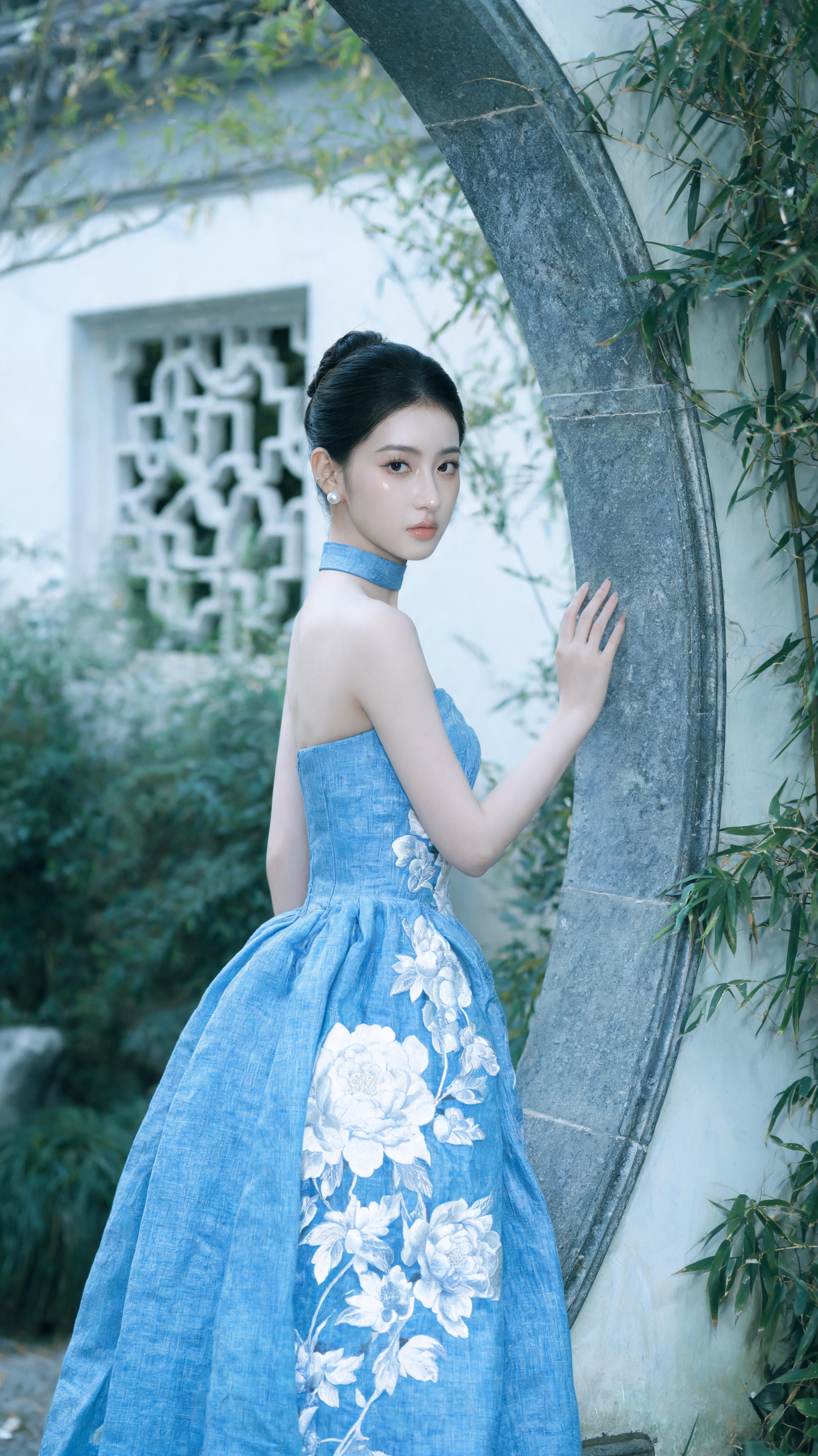 竖幅江南园林高级人像写真,年轻东亚女性,干净利落盘发,清冷优雅气质,小巧珍珠耳饰,侧身站立,单手轻扶灰色石材月洞门框,淡然看向镜头,蓝色抹胸长款礼服,同色系蓝色颈圈,裙身立体白色大朵牡丹刺绣,亚麻肌理面料,宽松花苞裙廓… - AI提示词参考图,来自西西提示词