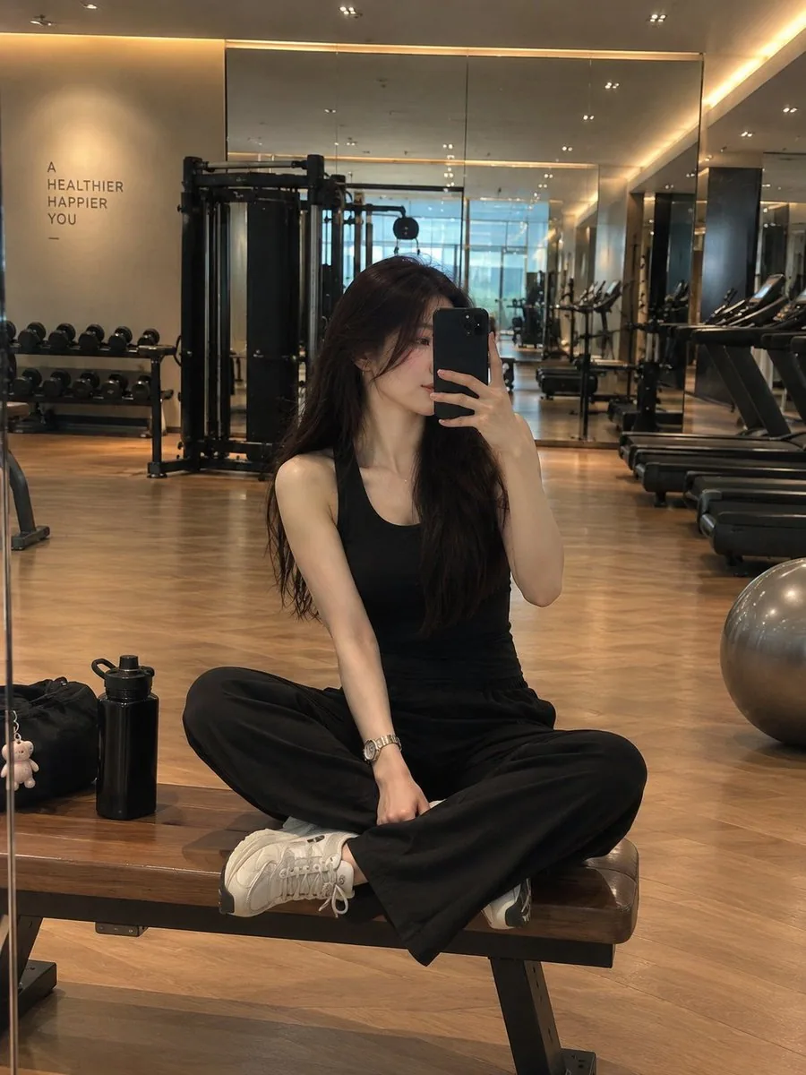 A stylish young woman taking a mirror selfie in a modern luxury gym, sitting cross-legged on a wooden workout… - AI提示词参考图，来自西西提示词